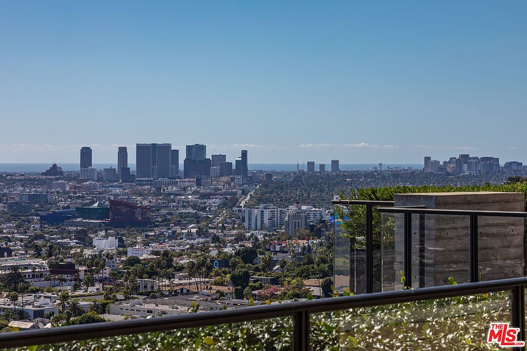 This image showcases a stunning panoramic view of a cityscape from a patio or balcony. The foreground features a modern railing and lush greenery, leading to a clear view of the city skyline under a bright blue sky. The overall impression is one of luxury and expansive living, highlighting the property's desirable location and outdoor living space.