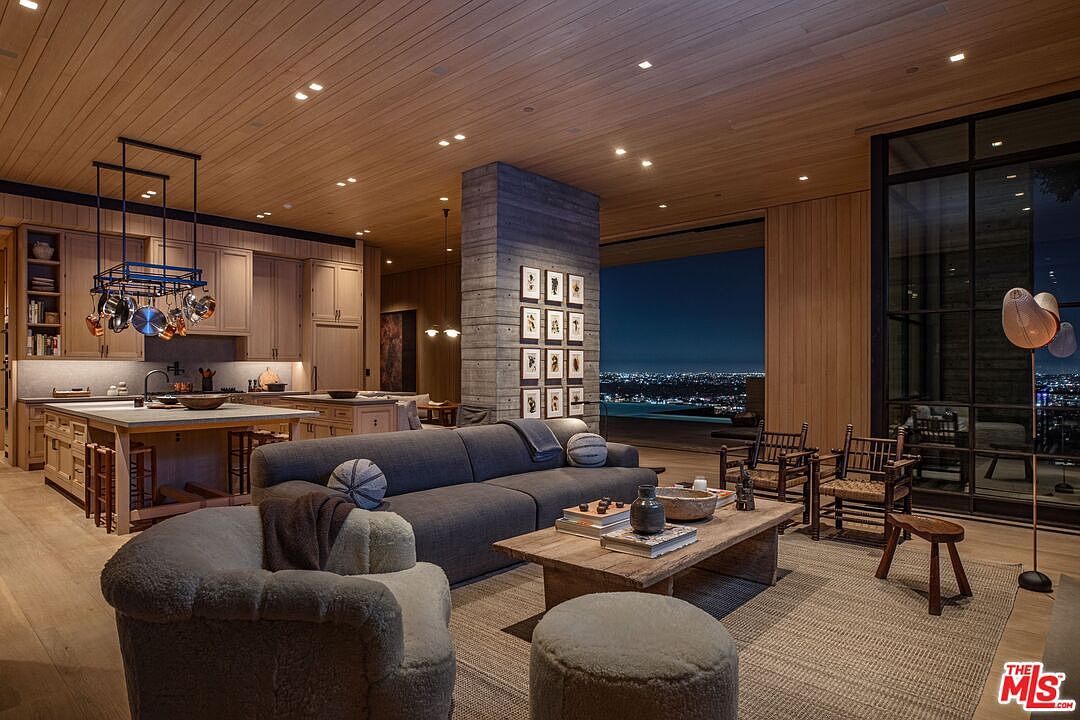 This expansive living room seamlessly blends with the kitchen area, featuring a modern aesthetic with wood-paneled ceilings and walls. A large gray sofa anchors the space, complemented by a rustic wooden coffee table and unique seating options. The room opens to a stunning city view through large windows, enhancing the luxurious and inviting atmosphere.