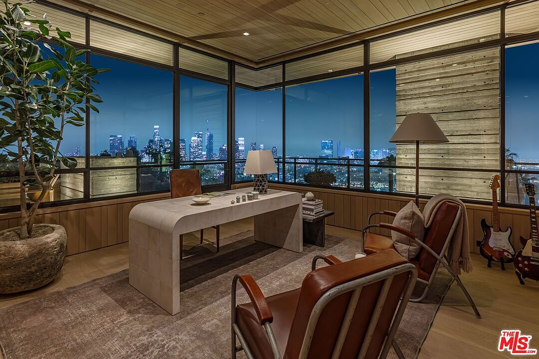 This interior shot showcases a modern office space with floor-to-ceiling windows offering a stunning city view at night. The room features a unique desk, comfortable leather chairs, and stylish lighting, creating a sophisticated and productive atmosphere. The wood paneling and natural elements, like the potted plant, add warmth and character to the space.