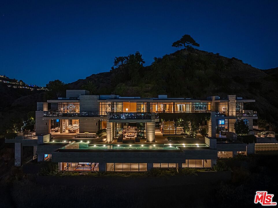 This aerial view showcases a luxurious, multi-level modern home nestled into a hillside at night. The property features extensive use of glass, allowing for illuminated interiors to be visible, and a sleek infinity pool with lighting. The surrounding landscape is a mix of natural terrain and manicured greenery, creating a sense of privacy and exclusivity.