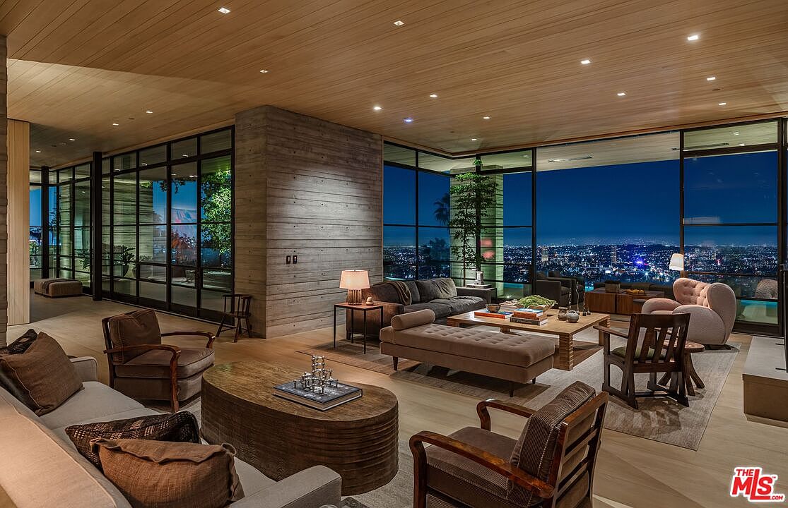 This is a luxurious living room featuring floor-to-ceiling windows that offer a stunning city view at night. The room is furnished with multiple seating areas, including sofas, armchairs, and a chaise lounge, arranged around wooden coffee tables. The interior design incorporates natural materials like wood and concrete, creating a modern and sophisticated atmosphere.