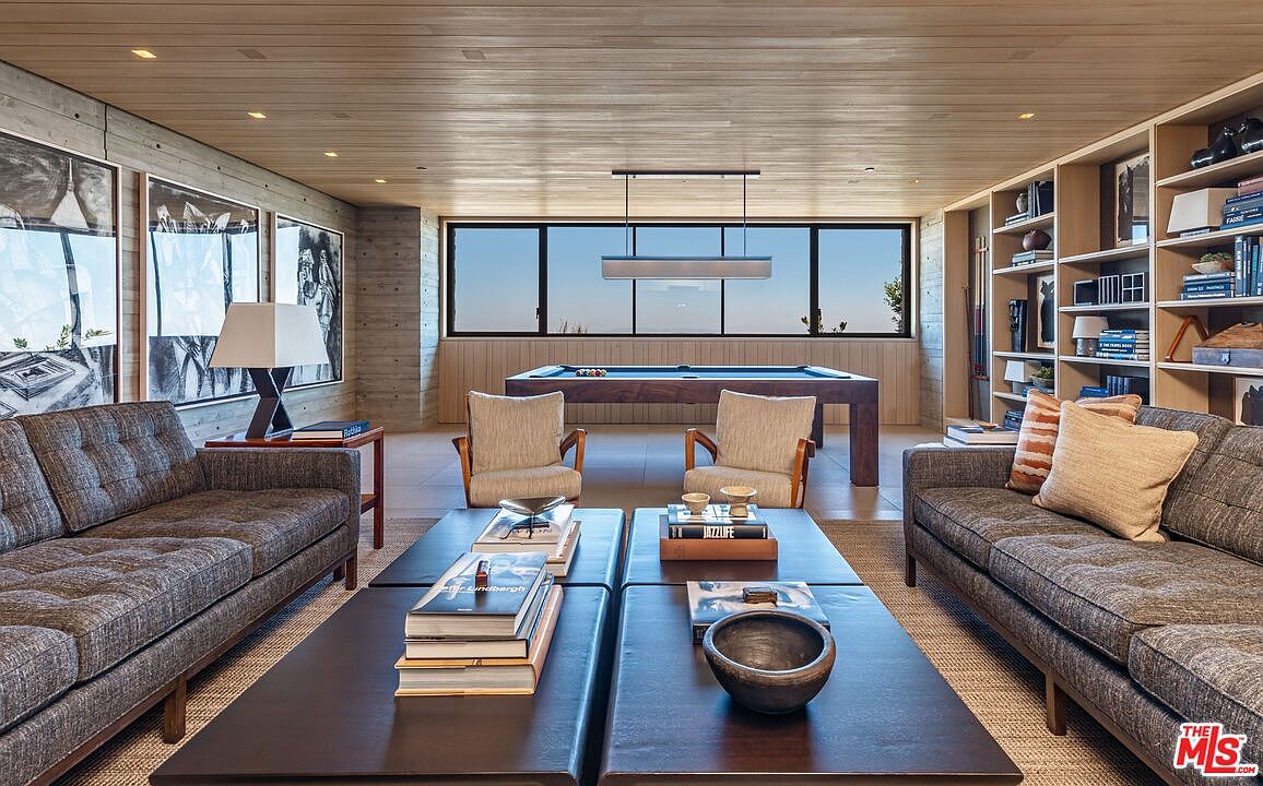 This is an interior shot of a modern living room featuring two gray sofas facing each other, separated by two dark-stained coffee tables. In the background, there is a pool table under a large window, and built-in bookshelves line the right wall. The room has a sophisticated and comfortable feel, with a focus on natural light and clean lines.