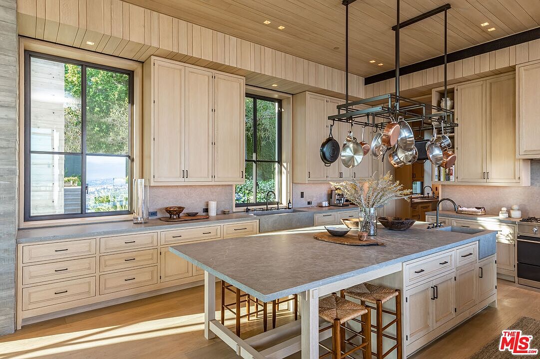 This is a bright and spacious kitchen featuring light wood cabinetry, a large center island with seating, and modern appliances. A pot rack hangs above the island, adding a touch of rustic charm. Large windows provide natural light and views of the outdoors, creating a warm and inviting atmosphere.