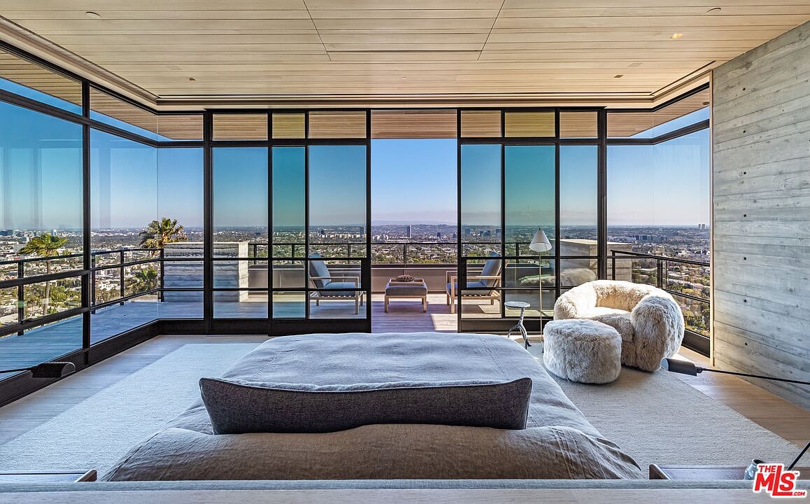 This primary bedroom boasts floor-to-ceiling windows offering panoramic city views. The room features a large bed with a neutral color palette, a cozy armchair with an ottoman, and a minimalist design. The space is bright, airy, and exudes a sense of luxury and tranquility.