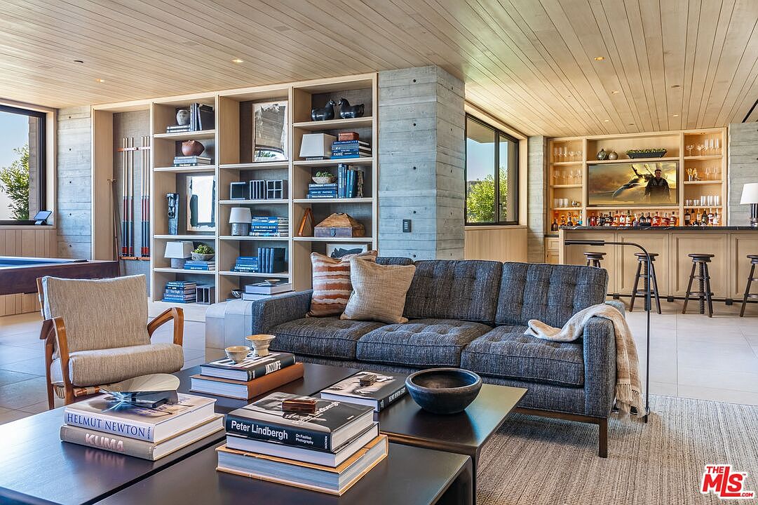 This is an interior shot of a modern living room featuring a gray sofa with throw pillows and a blanket, a dark coffee table with books, and a beige armchair. A large built-in bookshelf and a bar area with stools are visible in the background. The room has a contemporary design with a focus on natural materials and neutral colors.