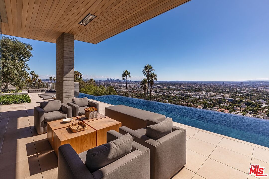 This image showcases a luxurious outdoor patio area with comfortable gray armchairs arranged around a modern wooden coffee table. An infinity pool stretches along the edge, offering stunning panoramic views of the city skyline and lush greenery. The space is designed for relaxation and entertainment, blending seamlessly with the natural surroundings.