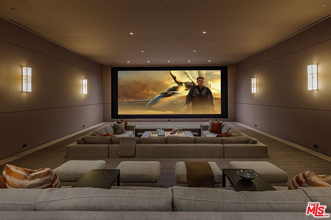 This is a luxurious home theater featuring a large screen displaying a scene from "Top Gun: Maverick". The room is designed with a large sectional sofa, ottomans, and side tables, providing ample seating. Wall sconces provide soft lighting, creating a cinematic atmosphere perfect for movie viewing.