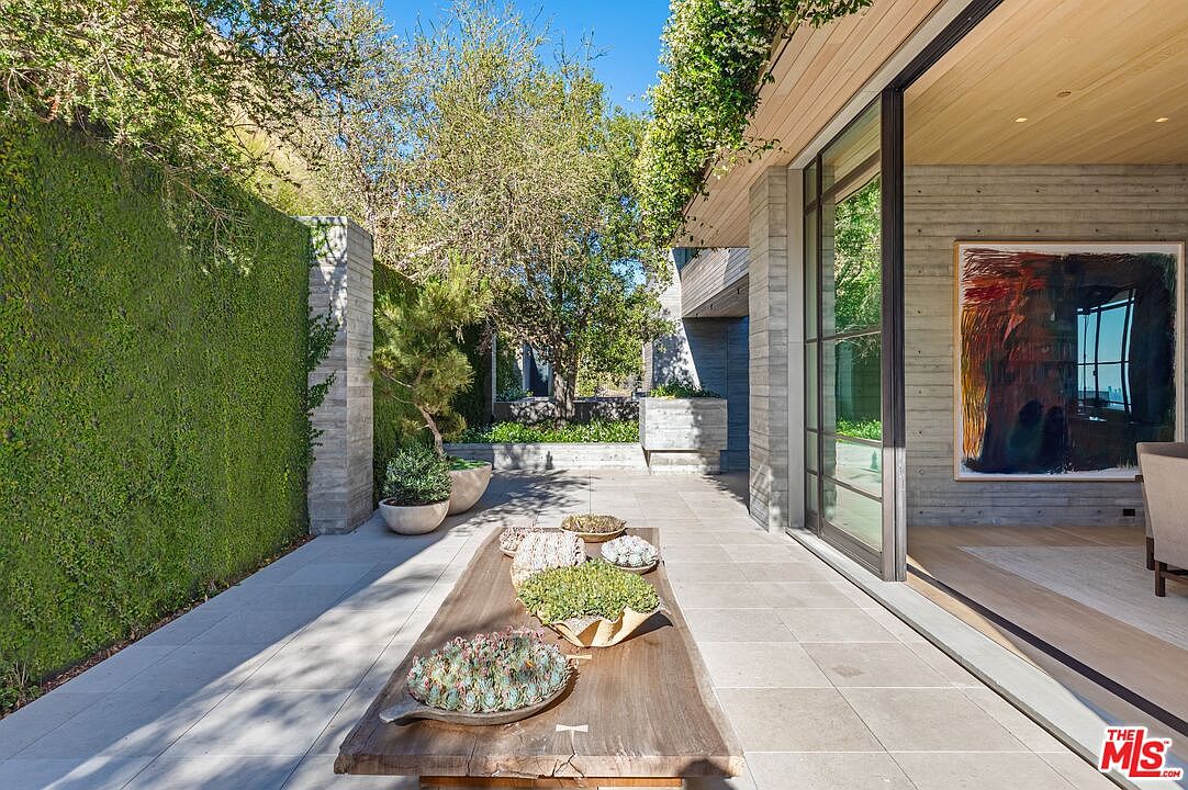 This image showcases a modern patio area with a long wooden table adorned with succulent arrangements, set against a backdrop of a lush green wall and contemporary architecture. The patio features large format tiles, and the adjacent building has expansive glass doors, creating a seamless indoor-outdoor transition. The overall impression is one of sophisticated tranquility and connection with nature.