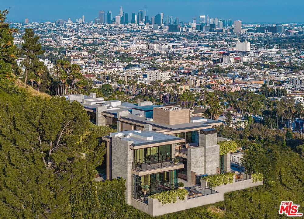 This aerial view showcases a modern, multi-level home nestled into a hillside, offering stunning panoramic views of the city skyline. The architectural design features clean lines, concrete elements, and expansive glass windows, blending seamlessly with the surrounding lush greenery. The property includes multiple outdoor terraces and balconies, perfect for enjoying the breathtaking scenery.