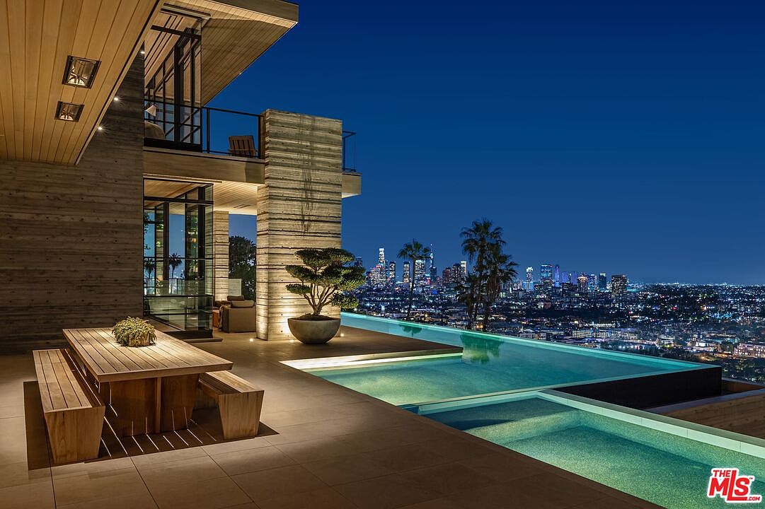 This image showcases a luxurious outdoor space featuring a modern infinity pool with stunning city views at night. The pool area includes a dining table with benches, a bonsai tree, and sleek architectural elements with wood and concrete finishes. The overall impression is one of high-end living and sophisticated design.