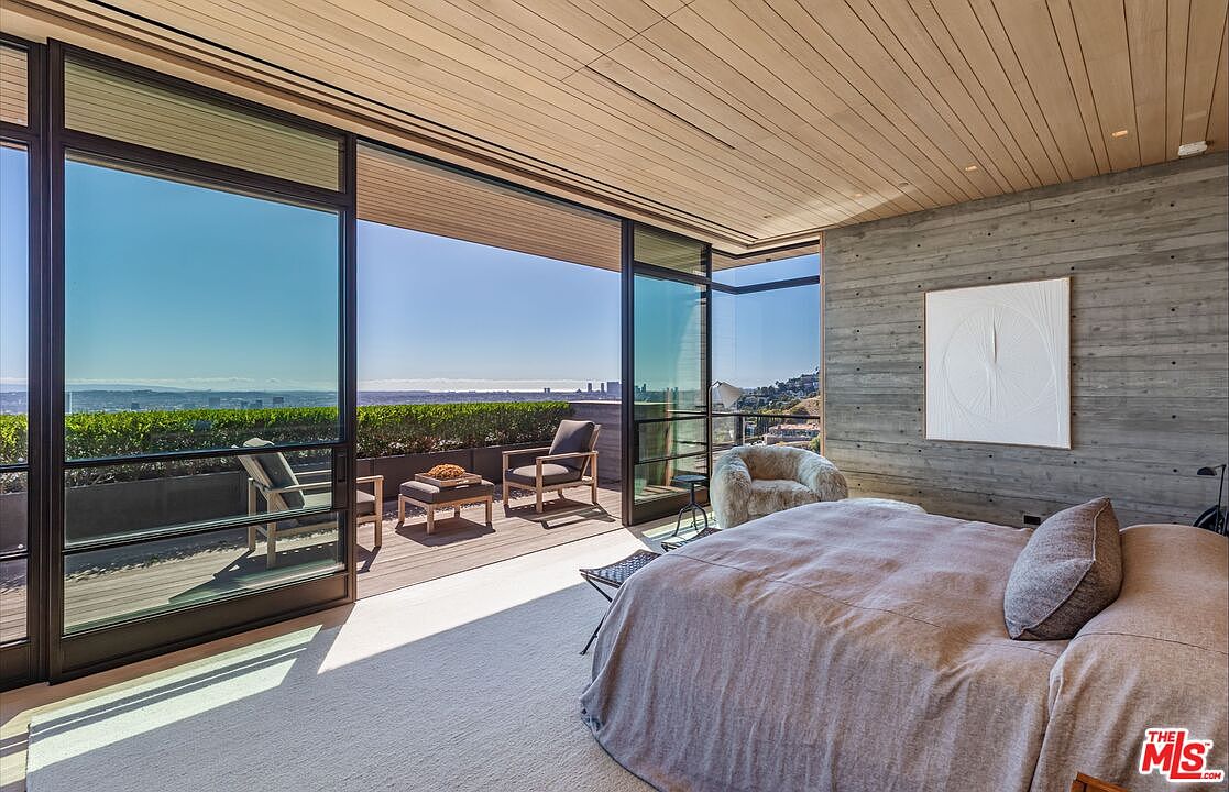 This is a luxurious primary bedroom featuring a modern design with a concrete accent wall and a light wood plank ceiling. Large sliding glass doors open to a private balcony with outdoor seating and a view of the city skyline. The room is well-lit with natural light, creating a serene and inviting atmosphere.