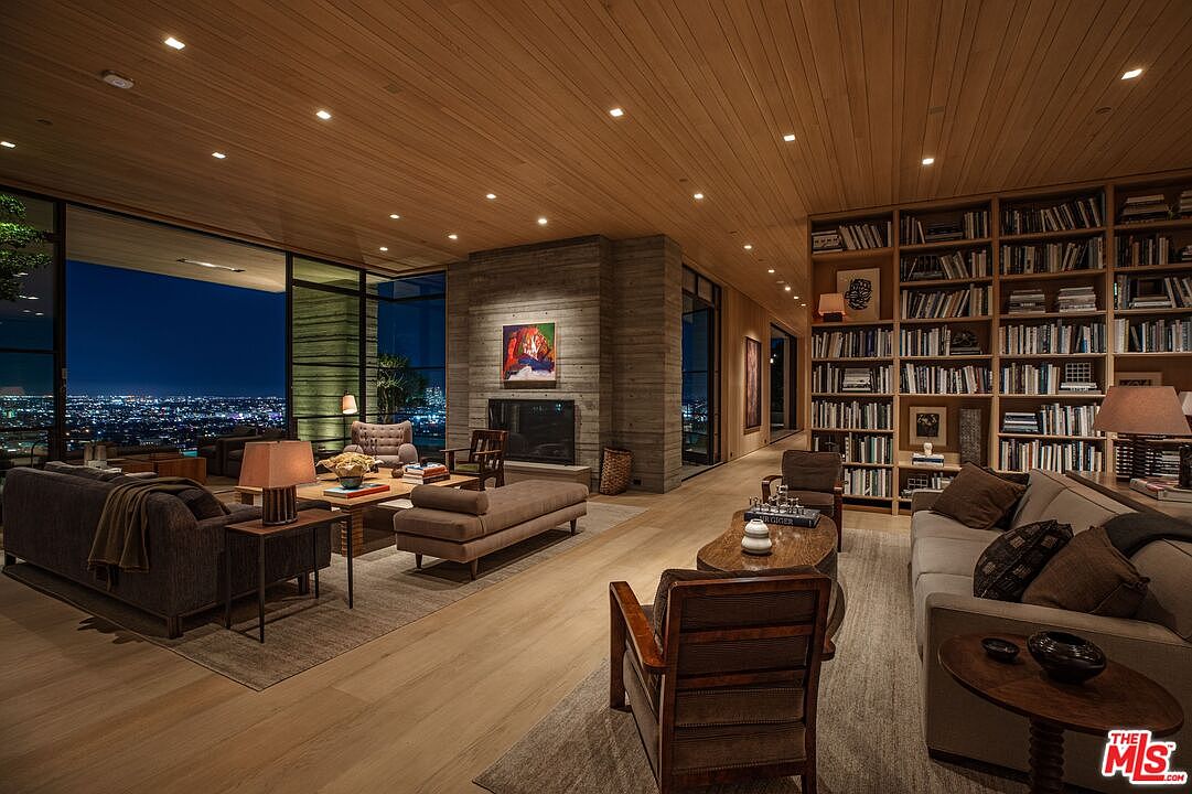 This is a luxurious living room featuring a floor-to-ceiling bookcase, a modern fireplace with an art piece above it, and expansive windows offering a stunning city view at night. The room is furnished with comfortable sofas, wooden chairs, and stylish coffee tables, creating a warm and inviting atmosphere. The wooden ceiling and recessed lighting add to the room's elegance and sophistication.