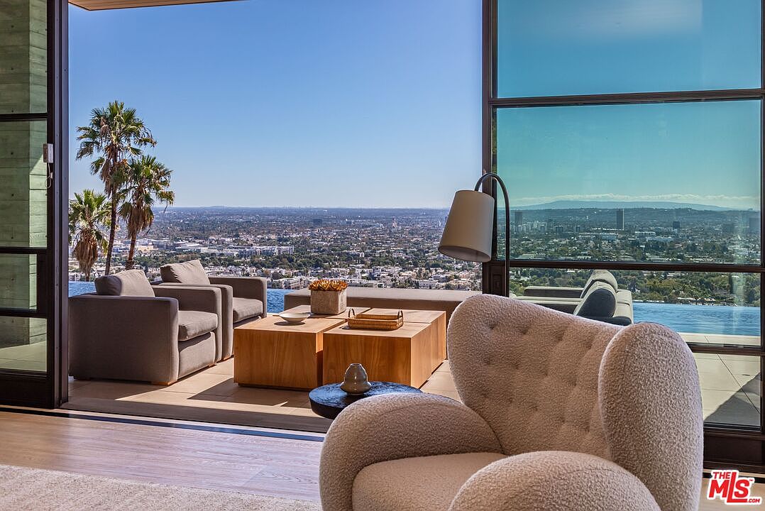 This image showcases a luxurious living room with an open-air design, featuring comfortable armchairs and stylish wooden coffee tables. The room seamlessly blends indoor and outdoor spaces, offering breathtaking panoramic views of the city skyline and palm trees. The design emphasizes modern elegance and relaxation, perfect for enjoying the scenic surroundings.
