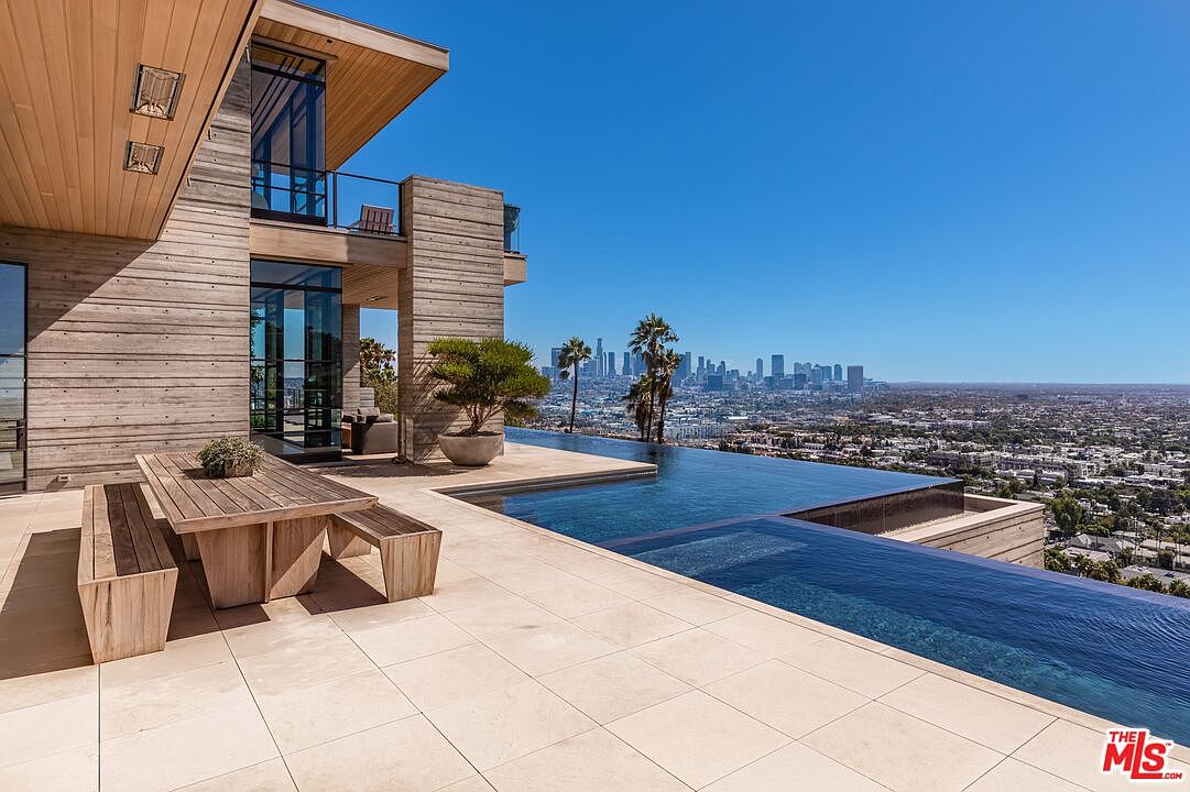 This image showcases a luxurious infinity pool overlooking a sprawling cityscape. The pool is surrounded by a modern patio with a wooden dining set, adjacent to a contemporary home with wood and glass architecture. The clear blue sky enhances the sense of opulence and relaxation, making it an ideal outdoor space for entertaining and enjoying panoramic views.