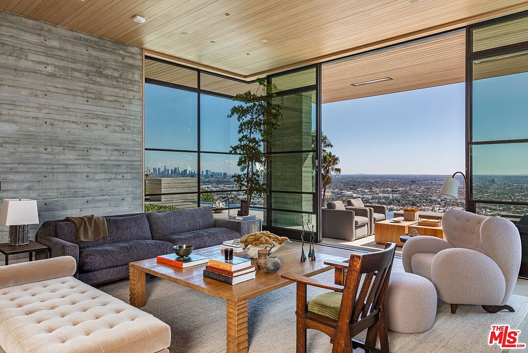 This is a luxurious living room featuring a modern design with floor-to-ceiling windows offering panoramic city views. The room is furnished with a plush gray sofa, a beige chaise lounge, and a unique wooden coffee table adorned with books and decorative items. An elegant armchair and ottoman complete the seating arrangement, all set on a neutral-toned rug, creating a sophisticated and inviting atmosphere.