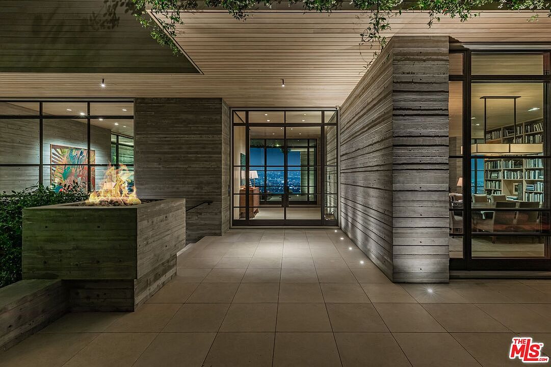 This image showcases a modern patio or deck area with a striking architectural design. The space features concrete walls with horizontal wood-like textures, large windows with black frames, and a fire pit. The flooring is composed of large square tiles, and the overall impression is one of sophisticated outdoor living with a view of the city lights in the background.