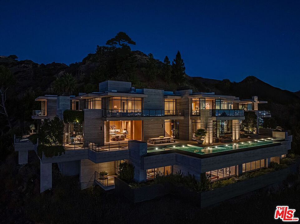 This aerial view showcases a modern, multi-level luxury home at night, featuring clean lines, large windows, and a flat roof. A striking infinity pool is illuminated, adding to the property's allure, while the surrounding landscape provides a sense of privacy and exclusivity. The architectural design emphasizes a seamless integration with the natural environment.