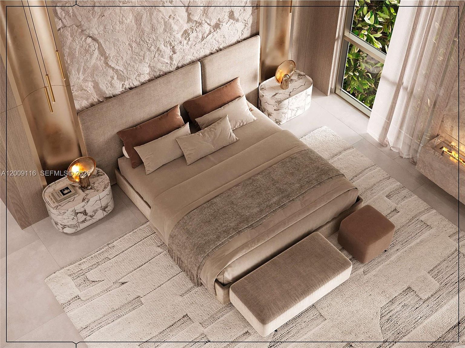 This high-angle shot captures a luxurious primary bedroom featuring a large bed with neutral-toned bedding and a textured, light-colored headboard set against a dramatic, rugged stone wall. The room is accented by two oval stone-topped nightstands with modern gold lamps and a plush, patterned area rug that anchors the space. A bench and ottoman at the foot of the bed, along with large windows overlooking greenery and a nearby fireplace, contribute to the sophisticated and serene atmosphere.