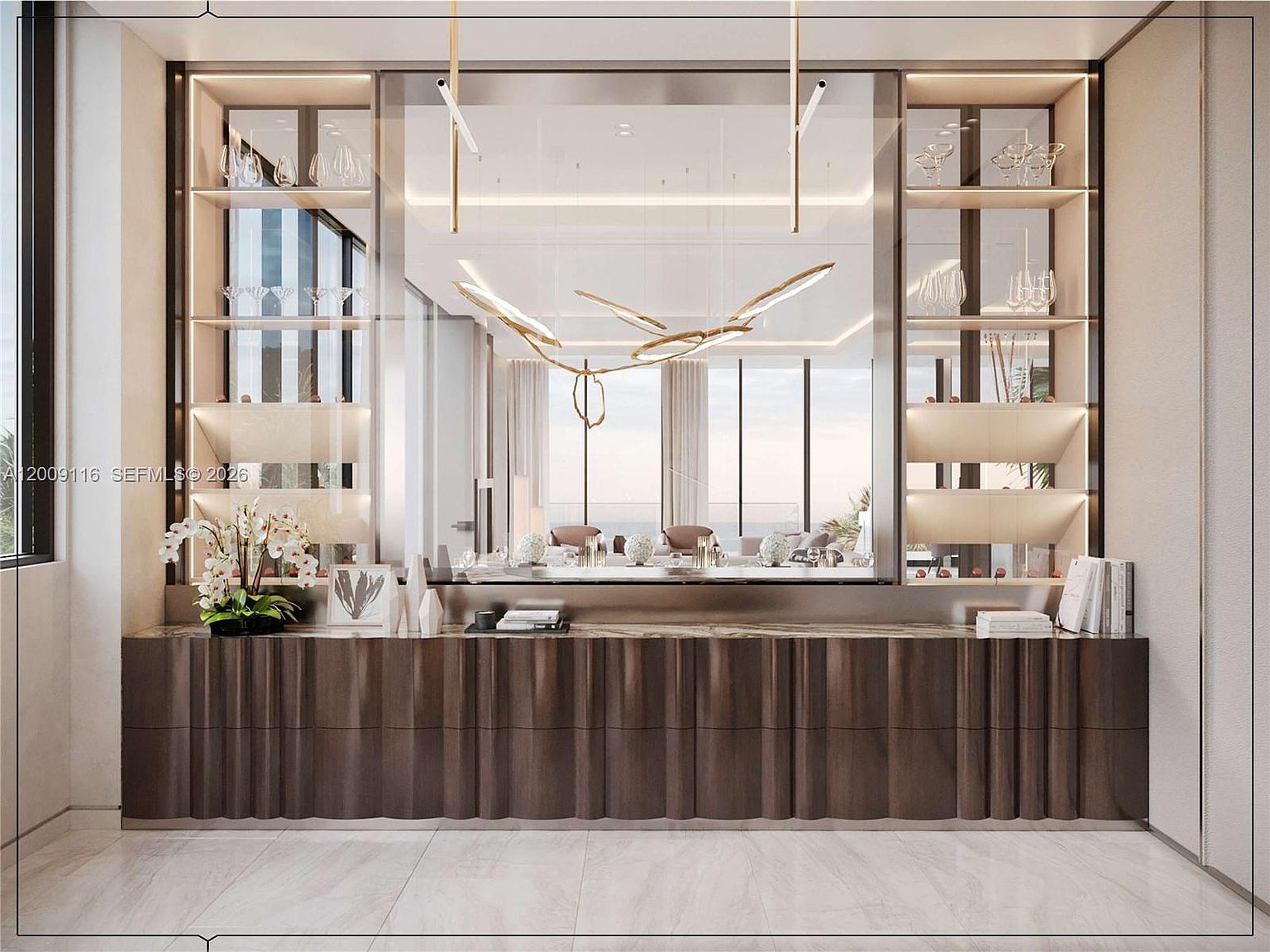 This sophisticated living area features a custom-built, dark-toned console with a textured, undulating front design positioned beneath a large, central mirror. Flanking the mirror are symmetrical, illuminated shelving units displaying glassware, while a sculptural, gold-toned light fixture hangs prominently in the foreground. The composition offers a refined, modern aesthetic with a clear view through the mirror into an adjacent living space with large windows overlooking the ocean.