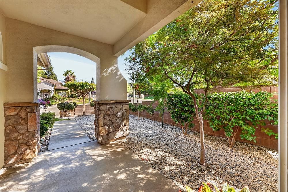 This image captures a covered entryway featuring stone-clad pillars that support a wide archway leading to a concrete walkway. The foreground shows a shaded porch area, while the background reveals a landscaped yard with gravel ground cover, mature trees, and a block wall perimeter. The perspective is from under the porch, looking out toward the street, creating a welcoming and sheltered transition space.