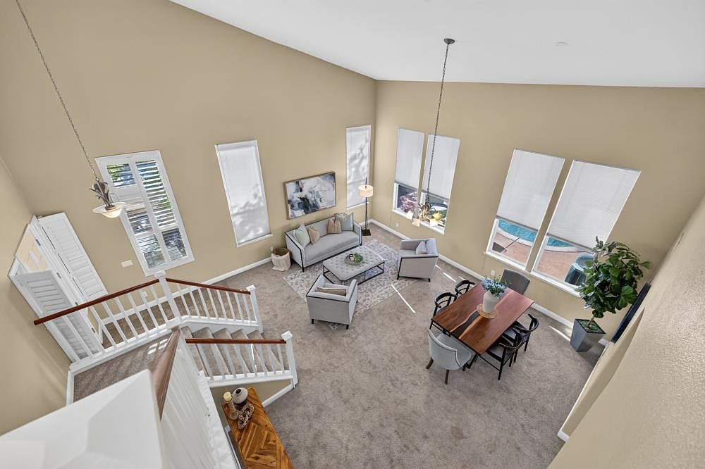This high-angle perspective captures an open-concept living and dining area characterized by soaring vaulted ceilings and neutral wall tones. The space features a comfortable seating arrangement with a light-colored sofa and armchairs centered around a patterned rug, alongside a formal dining area with a dark wood table. Large windows allow ample natural light to fill the room, highlighting the carpeted flooring and the staircase leading to the upper level.