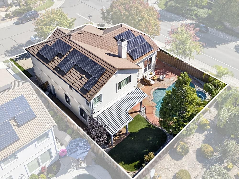This high-angle aerial view captures a suburban two-story home featuring a brown tile roof equipped with solar panels and a prominent chimney. The property includes a private backyard with a swimming pool, a covered patio area, and a small patch of green lawn, all enclosed by a wooden fence. The perspective provides a clear overview of the home's layout, its integration with the surrounding neighborhood, and the outdoor living space.
