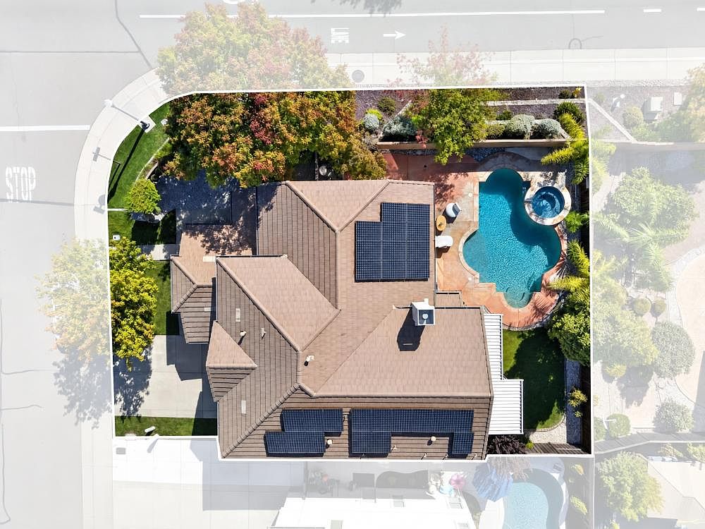 This high-angle aerial view captures a residential property featuring a brown-tiled roof equipped with solar panels. The backyard includes a uniquely shaped swimming pool with an integrated spa, surrounded by a paved patio area and mature landscaping. The property is situated on a corner lot, providing a clear perspective of the home's layout and its relationship to the surrounding neighborhood streets.