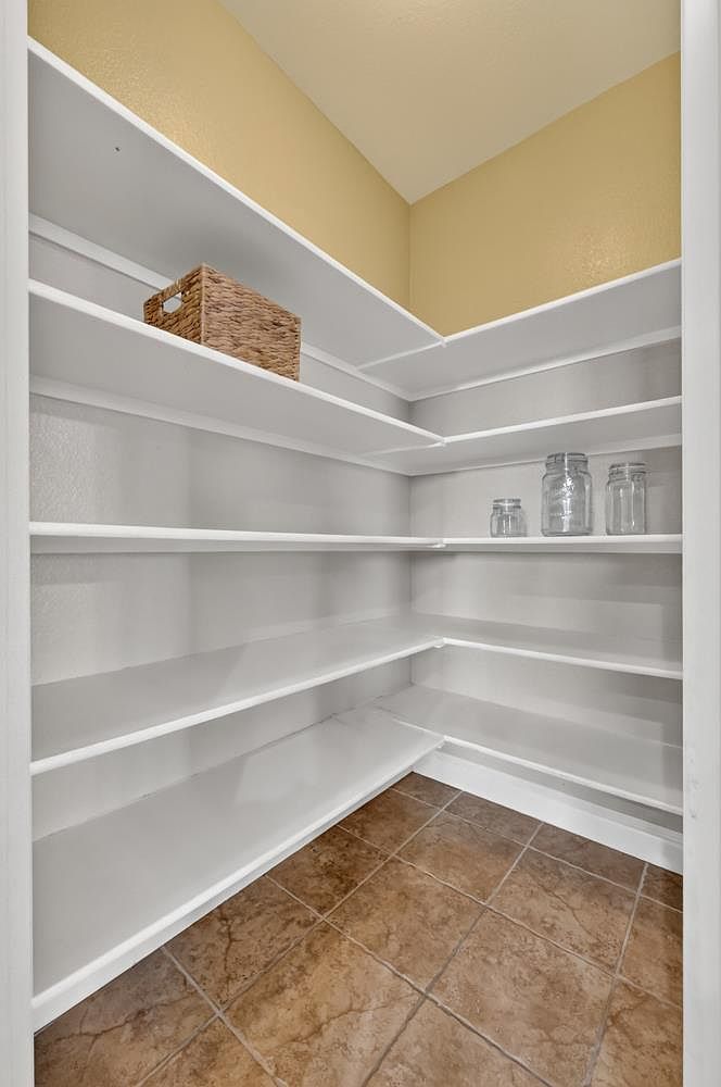 This walk-in pantry features L-shaped white shelving that provides ample storage space for kitchen essentials. The room is finished with neutral-toned walls and durable, earth-toned square floor tiles. A woven basket and a few glass jars are placed on the shelves, highlighting the functional organization of the space.
