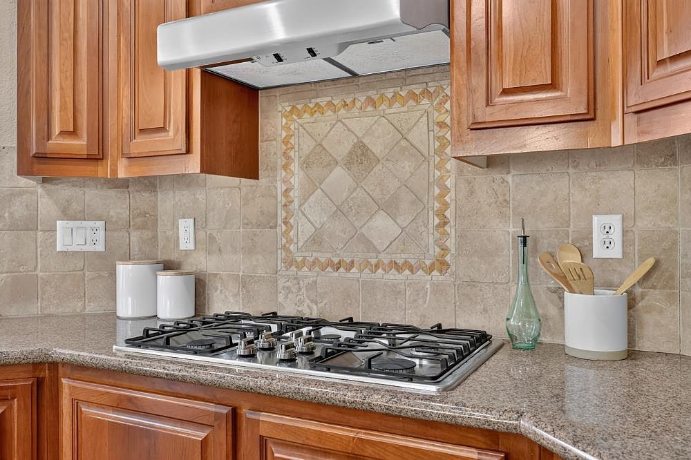This kitchen features warm-toned wood cabinetry paired with a speckled stone countertop and a neutral-toned tile backsplash. A stainless steel gas cooktop is centered beneath a matching range hood, which is accented by a decorative diamond-patterned tile inlay. The space is well-lit and includes practical storage containers and a utensil holder, creating a functional and inviting culinary environment.