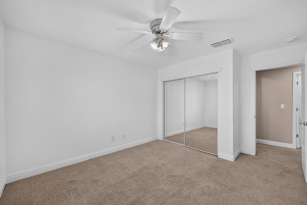 This bright, neutral-toned bedroom features plush wall-to-wall carpeting and a ceiling fan with integrated lighting. A large closet with mirrored sliding doors occupies one wall, reflecting the room and adding a sense of depth. The perspective is from the doorway, looking into the clean, minimalist space that leads toward an adjacent hallway.