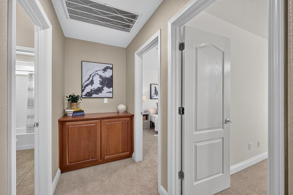 This interior hallway view features a neutral color palette with beige walls and light-toned carpeting. A wooden cabinet sits against the wall, topped with a small plant, books, and a decorative vase, while a framed abstract art piece hangs above it. The perspective looks through an open doorway into a bedroom, with a white door in the foreground leading to another room and a glimpse of a bathroom on the left.