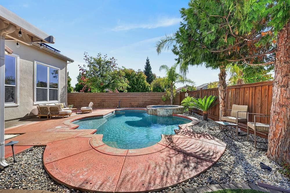 This backyard features a custom-shaped swimming pool with an integrated raised spa, surrounded by a stamped concrete deck in a warm terracotta hue. The area is landscaped with river rock beds, a mature tree, and a palm, all enclosed by a tall wooden privacy fence. Outdoor seating is arranged on the patio, creating a relaxing and private atmosphere perfect for entertaining.