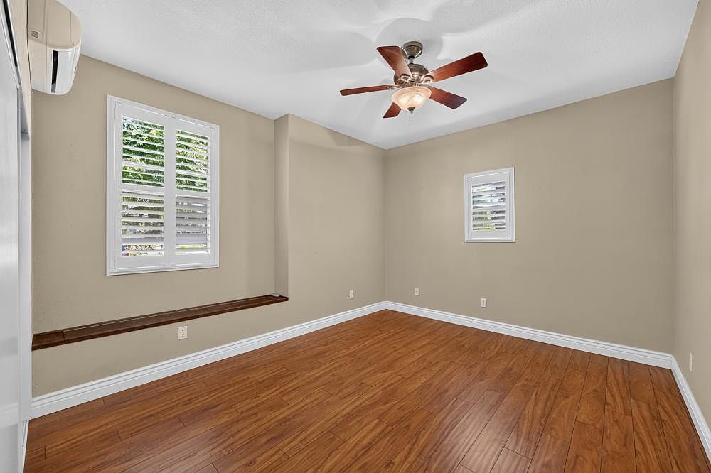 This bright, empty bedroom features warm-toned wood-look flooring and neutral beige walls, creating a versatile and inviting space. A ceiling fan with integrated lighting hangs centrally, while a window with white plantation shutters and a built-in wooden ledge adds architectural interest. The room is well-lit and provides a clean, blank canvas ready for personalization.