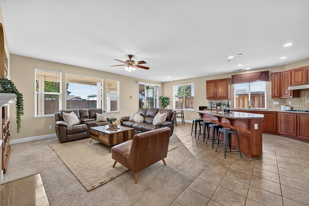 This open-concept living area features a spacious layout with a comfortable seating arrangement centered around a wooden coffee table. The room flows seamlessly into a kitchen with a large island and bar seating, creating an ideal space for entertaining. Large windows allow ample natural light to fill the space, highlighting the neutral color palette and the transition between the carpeted living area and the tiled kitchen floor.