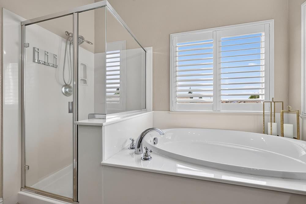 This primary bathroom features a clean, neutral aesthetic with a large soaking tub set into a white deck. Adjacent to the tub is a glass-enclosed shower stall with a handheld showerhead and a built-in niche. A window with white plantation shutters allows natural light to brighten the space, which is accented by decorative gold-toned lanterns on the tub deck.