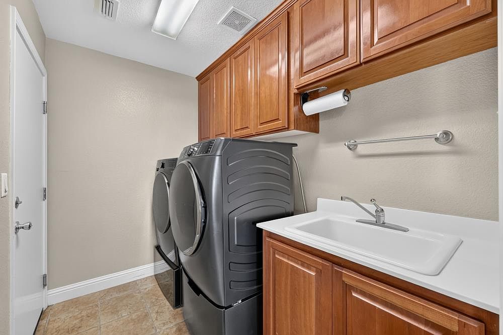 This functional laundry room features dark-toned, front-loading washer and dryer units positioned next to a utility sink with a white countertop and wood cabinetry. The space includes upper cabinets for storage, a wall-mounted towel bar, and a paper towel holder, all set against neutral-toned walls. The room is finished with tiled flooring and a white door, providing a clean and organized utility area.