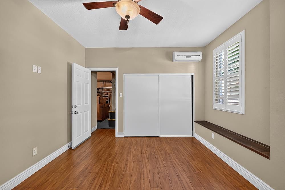 This clean, neutral-toned bedroom features warm wood-look flooring and a ceiling fan for climate control. A wall-mounted mini-split air conditioning unit is positioned above a set of white sliding closet doors, while a long, dark-toned built-in bench sits beneath a window with plantation shutters. The room offers a simple, functional layout with an open doorway leading to an adjacent storage area.