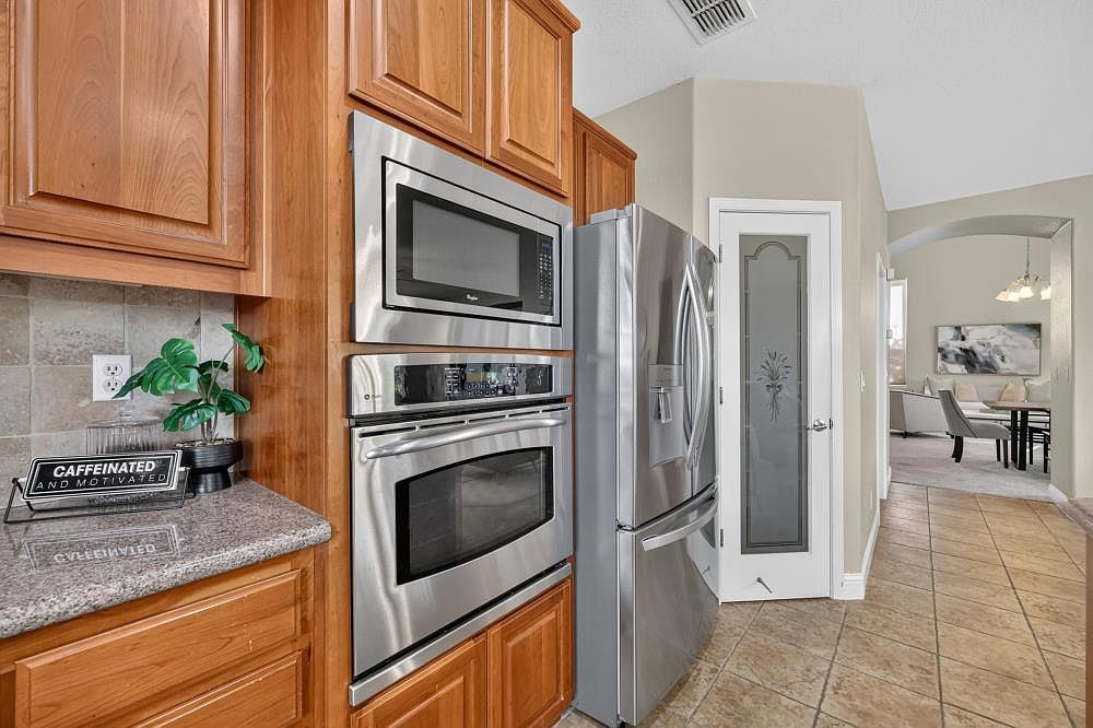 This kitchen features warm-toned wood cabinetry paired with stainless steel appliances, including a built-in microwave and wall oven. A granite countertop provides workspace, accented by a decorative sign and a small potted plant. The view transitions from the kitchen area into a bright, open-concept living space with tile flooring throughout.