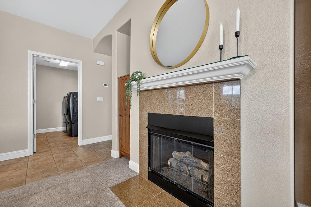 This interior view highlights a fireplace with a brown, speckled stone surround and a white mantel topped with two candle holders and a circular gold-framed mirror. To the left, a doorway leads into a laundry room containing a washer and dryer, while a tall, narrow wooden cabinet is tucked into the adjacent alcove. The room features a neutral color palette with a transition from carpeted flooring to tiled flooring near the fireplace hearth.