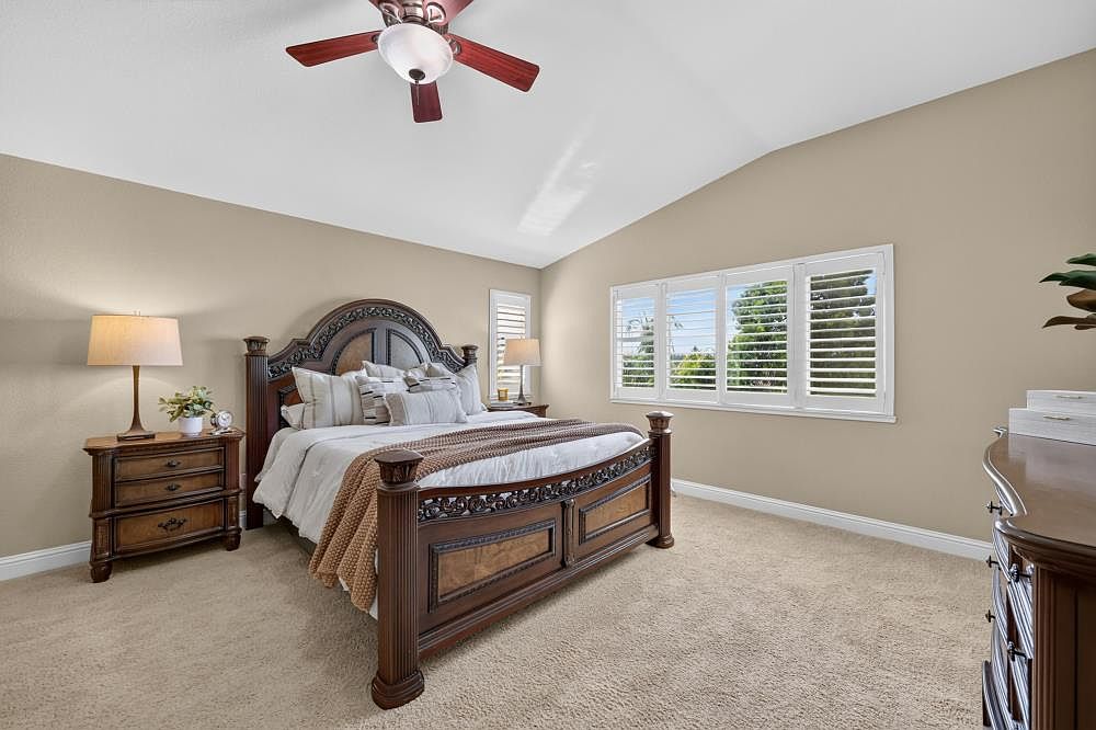 This spacious primary bedroom features a large, ornate wooden bed frame as the central focal point, complemented by matching nightstands and a dresser. The room is bathed in natural light from a wide set of windows adorned with white plantation shutters, creating a bright and airy atmosphere. Neutral wall tones and plush carpeting provide a warm, inviting foundation for this well-appointed retreat.