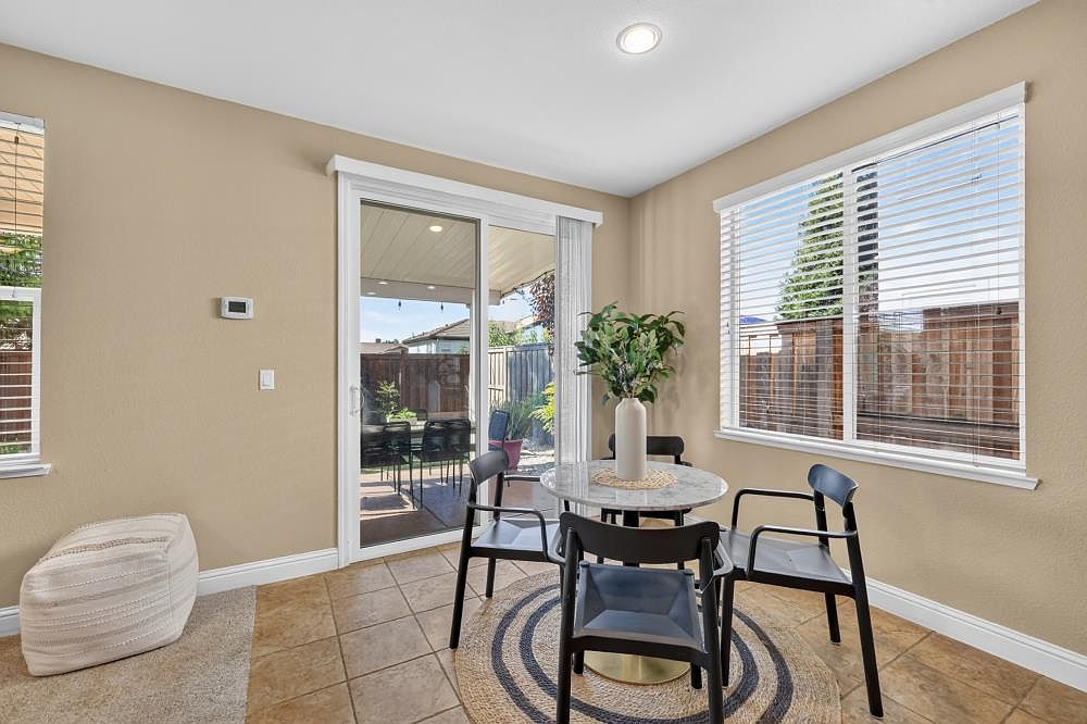 This bright dining area features a small, round table with a stone-look top and four modern black chairs, set atop a circular woven rug. Large sliding glass doors provide a clear view of the backyard patio, while a nearby window with horizontal blinds allows for ample natural light. The space is finished with neutral-toned walls and tiled flooring, creating a clean and inviting atmosphere.