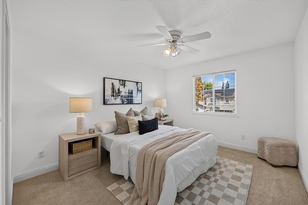 This bright and inviting bedroom features neutral walls and plush carpeting, creating a cozy atmosphere. The room is centered around a bed dressed in white linens with a textured throw blanket, flanked by two light wood nightstands topped with matching lamps. A framed piece of abstract art hangs above the headboard, and a window provides natural light while offering a view of the neighborhood.