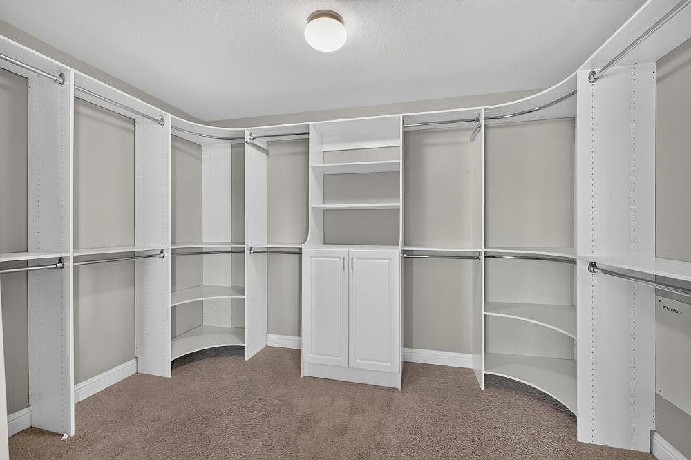 This spacious walk-in closet features custom white shelving and hanging rods arranged in a U-shape configuration. The center unit includes open shelving above a closed cabinet, providing versatile storage options, while curved corner shelves maximize the available space. The room is finished with neutral wall paint and plush carpeting, creating a clean and organized aesthetic.