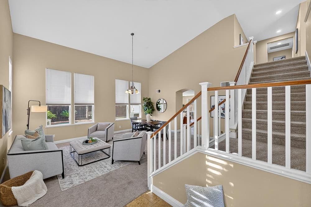 This bright and airy living room features high vaulted ceilings and a neutral color palette, creating an inviting open-concept atmosphere. The space is furnished with a light gray sofa, matching armchairs, and a central ottoman, all arranged on a patterned area rug. A staircase with a white railing leads to the upper level, while large windows allow for abundant natural light to fill the room.