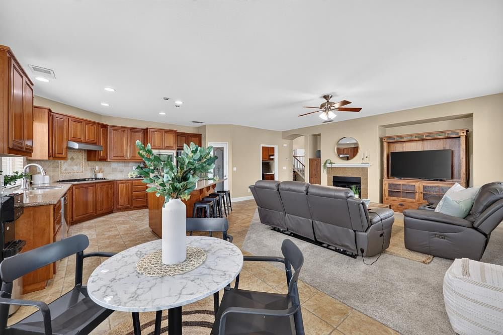 This open-concept living space features a kitchen with warm-toned cabinetry, stone countertops, and a central island with bar seating. The adjacent living area is anchored by a gray reclining sofa set and a built-in entertainment center with a television. A small, round dining table with a marble-look top sits in the foreground, creating a cohesive and functional flow between the cooking and relaxation zones.