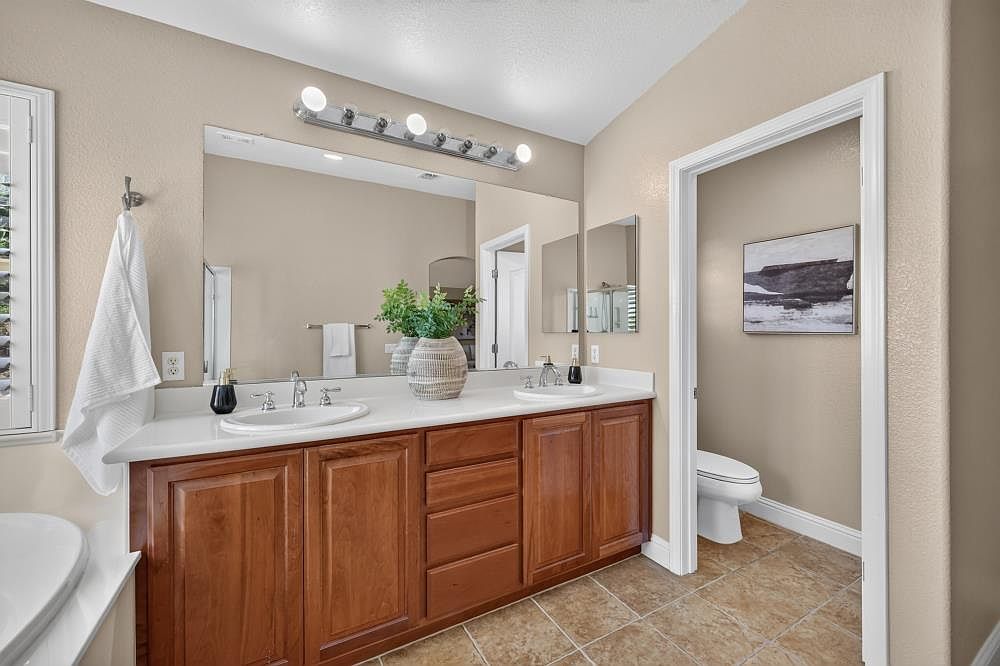 This primary bathroom features a double-sink vanity with warm-toned wood cabinetry and a white countertop. A large, full-width mirror reflects the space, while a light fixture with multiple bulbs provides bright illumination. To the right, a doorway leads into a private water closet area containing a toilet and wall art.