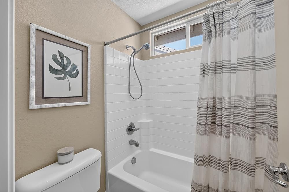 This compact bathroom features a white tub-shower combination with a subway-style tile surround and a handheld showerhead. A striped fabric shower curtain adds a touch of pattern, while a framed botanical print hangs on the neutral-toned wall above the toilet. The space is illuminated by natural light streaming through a high, horizontal window positioned above the shower area.