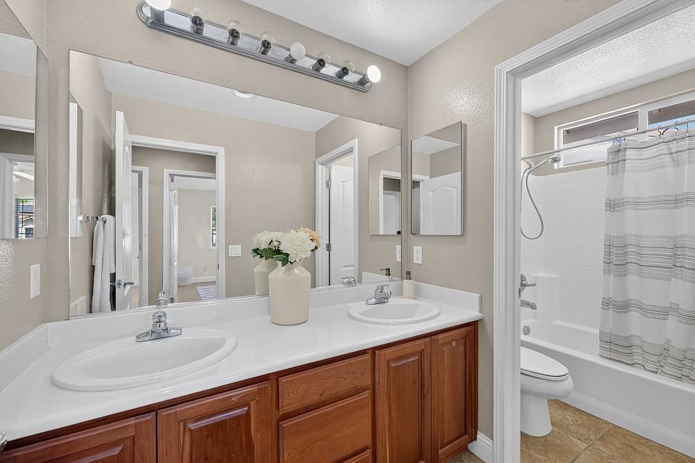 This bright, functional bathroom features a dual-sink vanity with warm-toned wood cabinetry and a white countertop. A large, full-width mirror reflects the space, while a separate shower-tub combination with a striped curtain is visible through the doorway. The neutral wall color and clean lines create a welcoming and well-maintained atmosphere.
