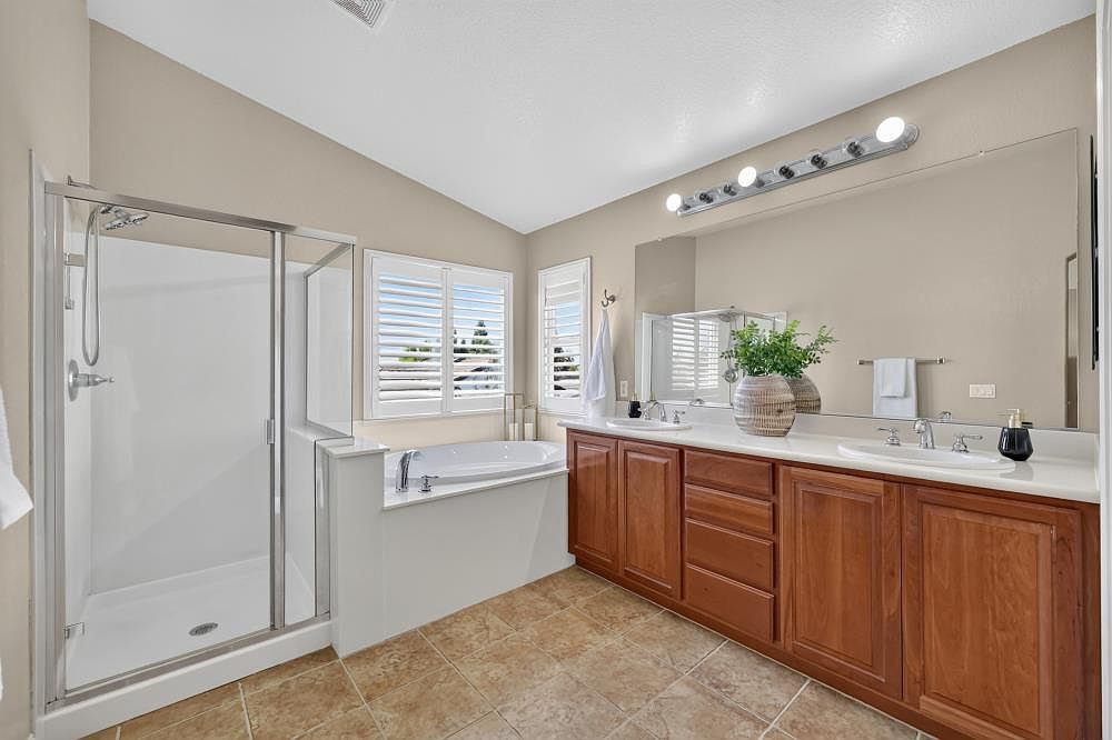 This spacious primary bathroom features a double-sink vanity with warm-toned wood cabinetry and a large wall-to-wall mirror. To the left, a glass-enclosed shower stall sits adjacent to a built-in soaking tub positioned beneath two windows with white plantation shutters. The room is finished with neutral-toned floor tiles and light-colored walls, creating a bright and functional atmosphere.