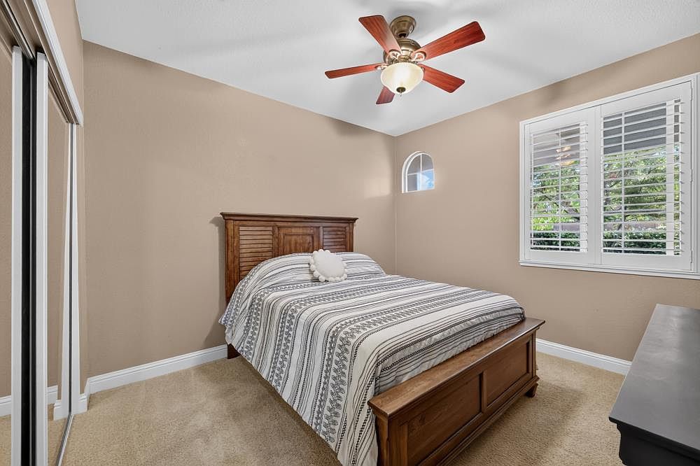 This cozy bedroom features a wooden bed frame with a slatted headboard, dressed in a neutral-toned striped comforter. The room is painted in a warm beige hue and includes a ceiling fan with a light fixture, a small arched window, and a large window covered by white plantation shutters. The space is carpeted and includes a sliding mirrored closet door on the left, creating a bright and inviting atmosphere.